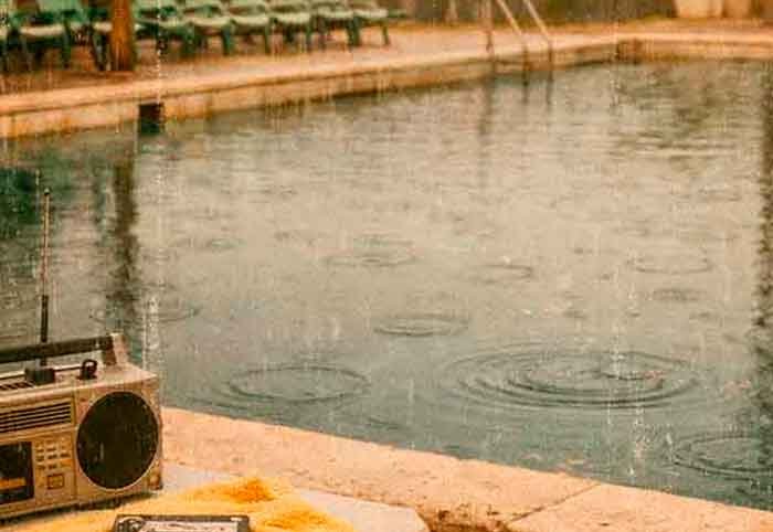 Piscina municipal vacía bajo la lluvia en Mallorca, con altavoces oxidados y círculos en el agua, evocando la melancolía de la canción Ciutat de Jaume Escala.