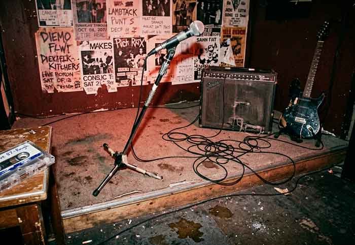 Escenario pequeño y desgastado con micrófono torcido, amplificador viejo y una cinta sin carátula, en una escena cruda y punk.