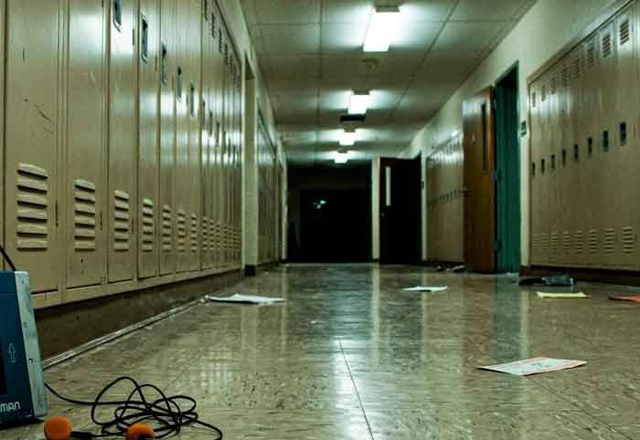 Pasillo largo y vacío de un instituto con un walkman apoyado en la pared, en una escena cruda y desaturada.