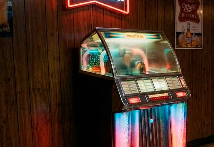 Máquina de música iluminada en un bar vacío de los 80, con un vaso y una moneda sobre la mesa, en una escena poética y melancólica.