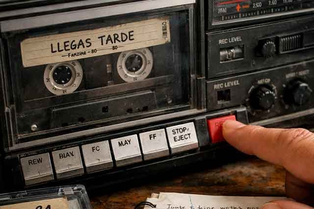 Mano presionando el botón REC de un radiocasete vintage para grabar música de la radio, con cintas y libreta sobre la mesa, evocando la nostalgia analógica de los años 80