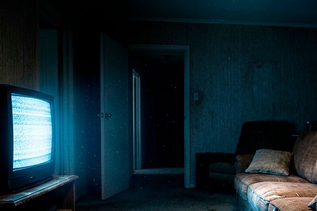 Salón oscuro iluminado solo por un televisor antiguo, con sofá marrón y objetos domésticos, en una escena cinematográfica y nocturna.