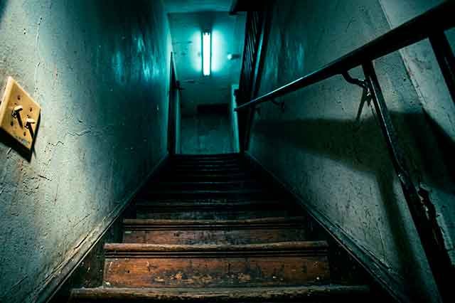 Escalera antigua iluminada por un fluorescente parpadeante, con sombras largas y ambiente cinematográfico frío.