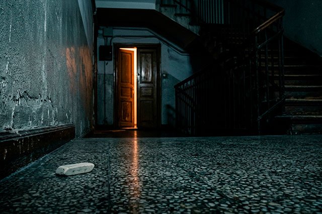 Rellano nocturno con suelo de terrazo, una goma de borrar en el suelo y una puerta entreabierta iluminando la penumbra.