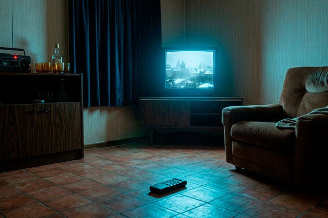 Salón nocturno iluminado solo por un televisor azul, con baldosas frías, un sillón hundido y un piloto rojo encendido en un radiocasete.