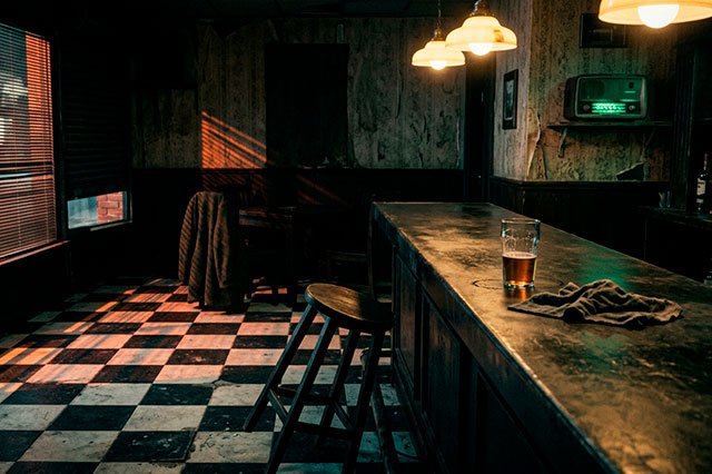 Bar nocturno con barra iluminada, azulejos hacia el baño, neón filtrado por la persiana y una radio encendida en penumbra.