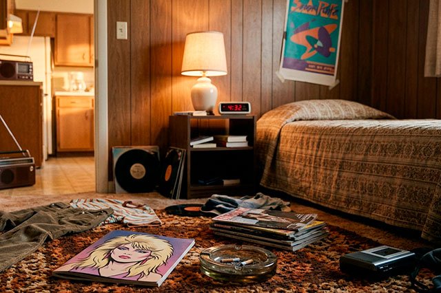 Dormitorio juvenil ochentero con una revista musical genérica abierta en el suelo junto a un cenicero y una cama individual al fondo