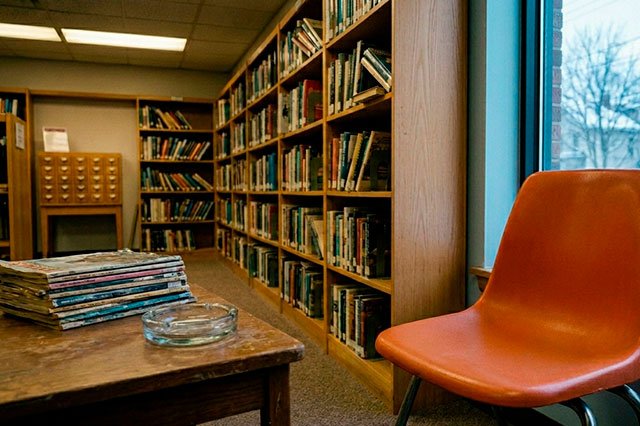 Silla de plástico naranja en una biblioteca ochentera con una revista genérica doblada sobre el asiento y luz fría entrando por la ventana