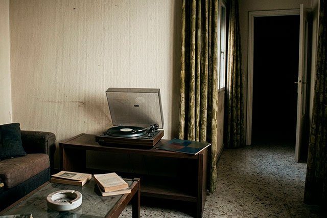 Salón modesto ochentero con un tocadiscos y un single genérico girando en silencio junto a un pasillo oscuro